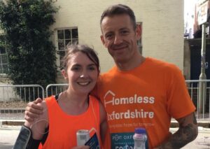 Amber and Chris stand smiling after finishing the Oxford Half. Both are wearing their orange Homeless Oxfordshire t-shirts