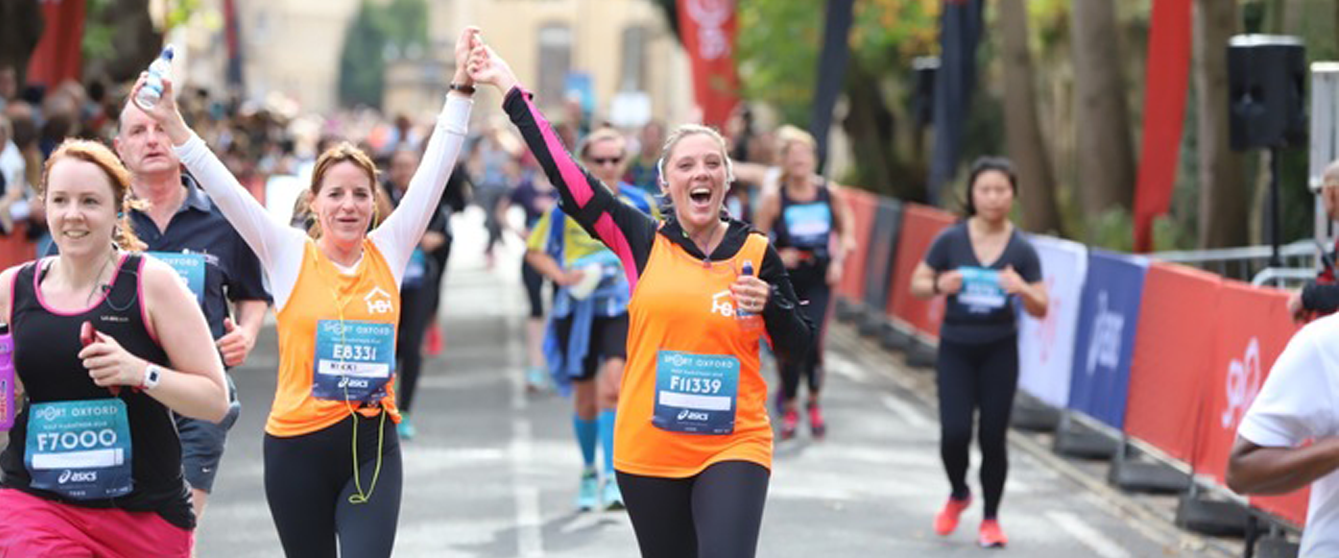 Nikki and Rachel cross the finish line hands up in the air, celebrating. Both are wearing their Homeless Oxfordshire orange running vests.